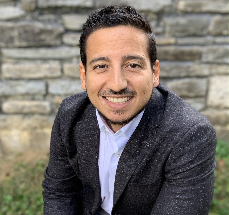Rene Gomez smiling outdoors in a charcoal jacket and white shirt.